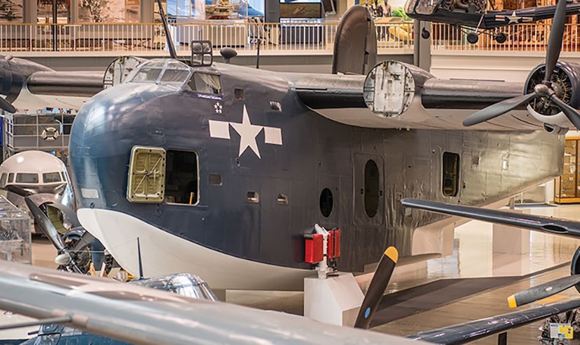 A large seaplane sits on the tiled floor of a museum. Other aircraft and their wings can be seen nearby.