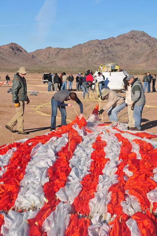 Four men and a woman all dressed in civilian clothing, pull an orange and white striped parachute from the ground. A group of people surround a space capsule in the background.