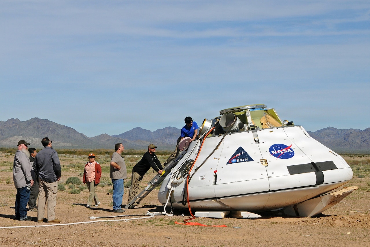 People surround a white space capsule as it sits on the ground. A desert mountain range is in the distant background.
