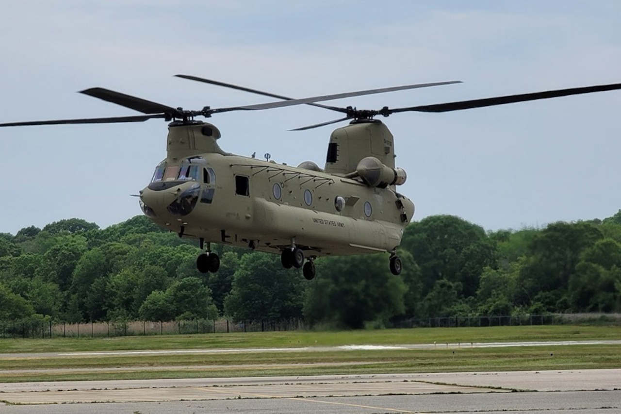 A large military helicopter hovers above a tarmac. There is a wooded area in the background.