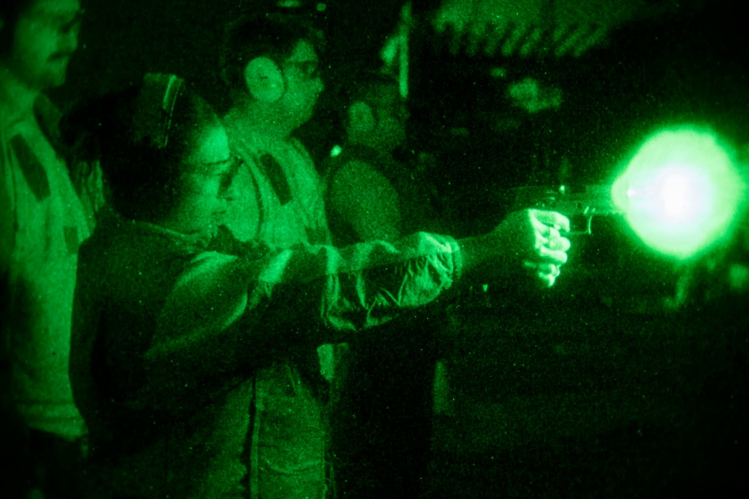 A woman in a dark area wears protective headphones and goggles while firing a weapon that produces a green and white flash from the nozzle; three men, also wearing protective headphones and goggles, stand to the left and observe.