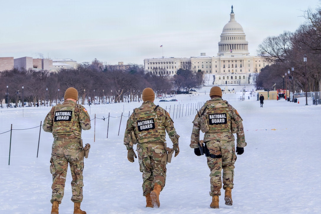 Three people wearing camouflage military uniforms and covers walk in the snow, side by side, toward the U.S. Capitol. Several people and trees are in the background.