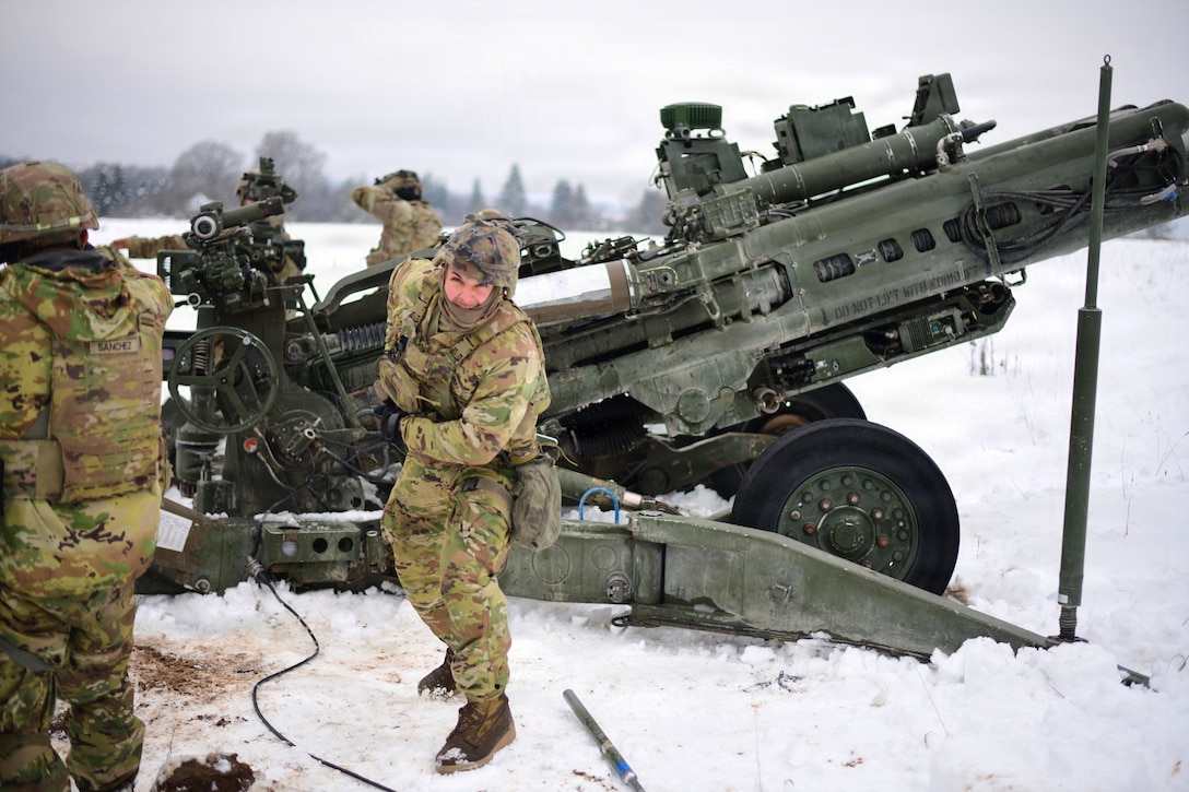 Four people wearing camouflage military uniforms and cold-weather attire stand outside in the snow, working on a large military weapon system during daytime, with trees in the distance.