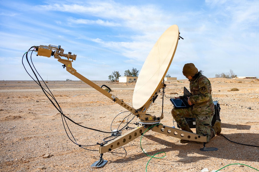 A person wearing a camouflage military uniform and cover kneels in desert terrain while holding a laptop connected to a large satellite, outside under a blue sky.