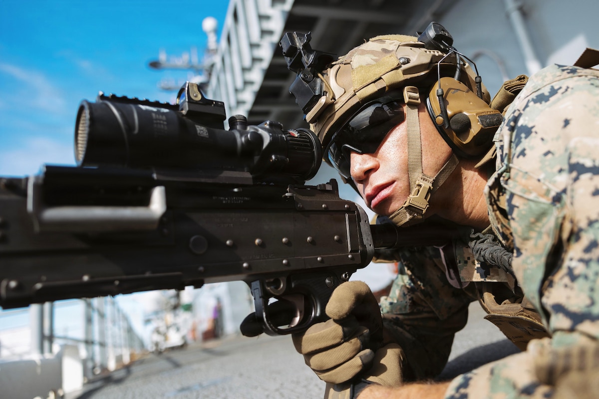 A man wearing a camouflage military uniform, helmet, protective sunglasses and gloves lies on his stomach to look through the scope of the weapon he is holding.