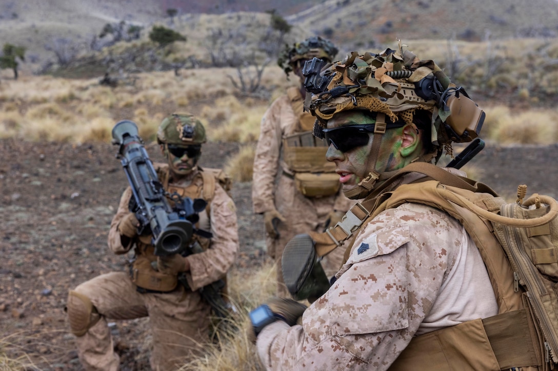 Two men wearing camouflage military uniforms, face paint and helmets kneel in rocky terrain; one holds a large weapon while the other looks into the distance. Another partially visible person stands to the left looking on.