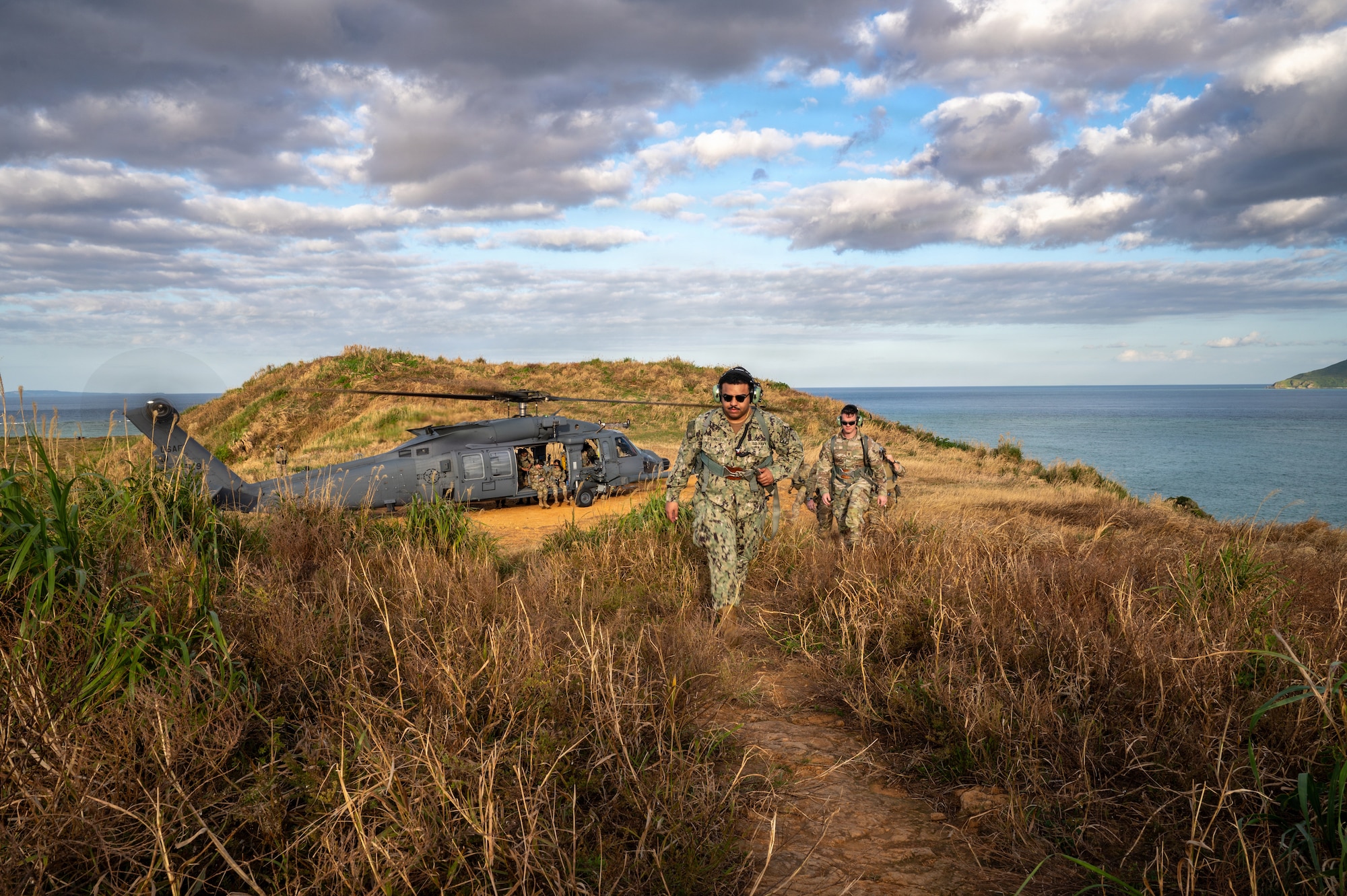 Two service members walk towards the camera, behind them is an HH-60W Jolly Green II helicopter.