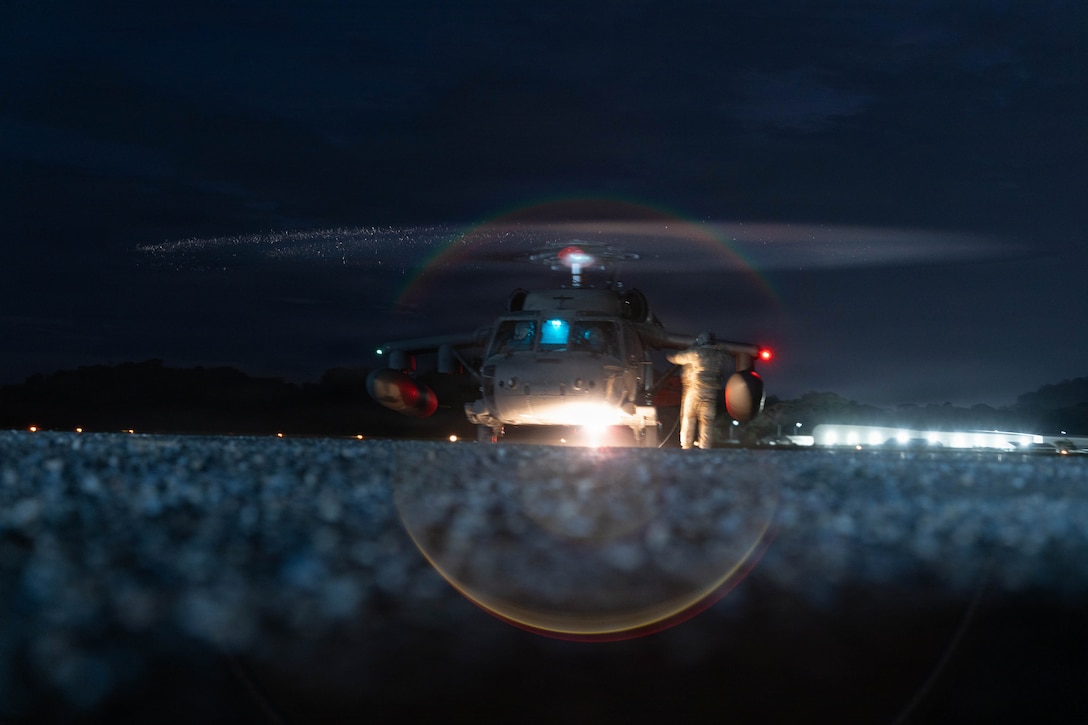 A soldier stands besides a military helicopter at night.