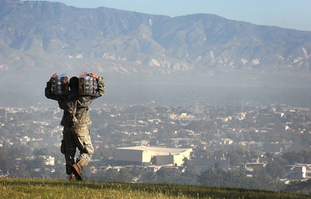 A soldier is walking away from the camera with cases of water on his back