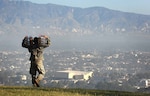 A soldier is walking away from the camera with cases of water on his back