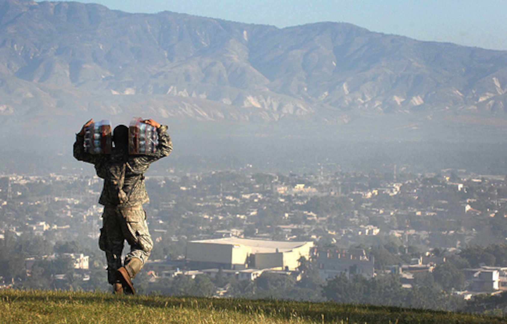 A soldier is walking away from the camera with cases of water on his back