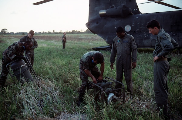 Soldiers stand in tall grass next to a Chinook helicopter