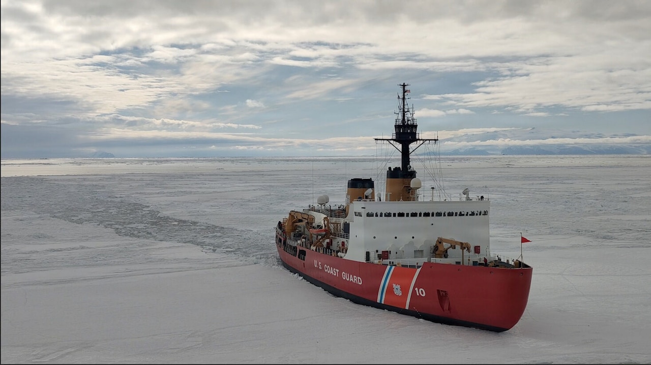 A large ship with the words, “U.S. Coast Guard” written on the side, sails through icy water, leaving a trail of broken ice and open water behind it.