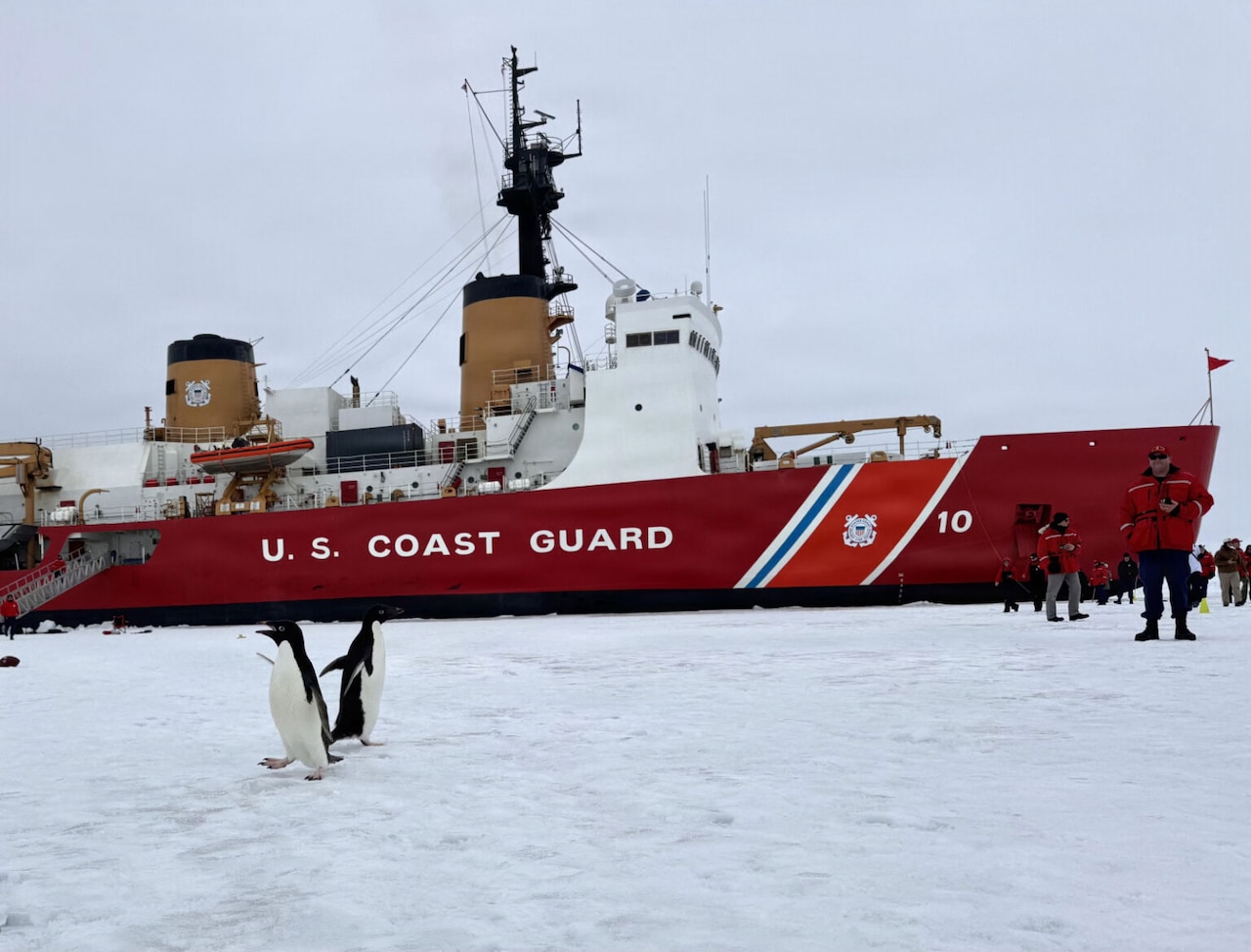 Dozens of people in winter attire and two penguins walk on ice as a large ship is docked in the background with the words, “U.S. Coast Guard,” written on the side.