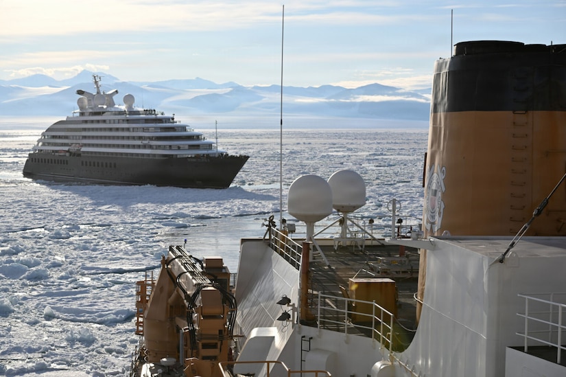 A large ship sails through icy water as a cruise ship follows close behind. There are snow-covered mountains in the background.