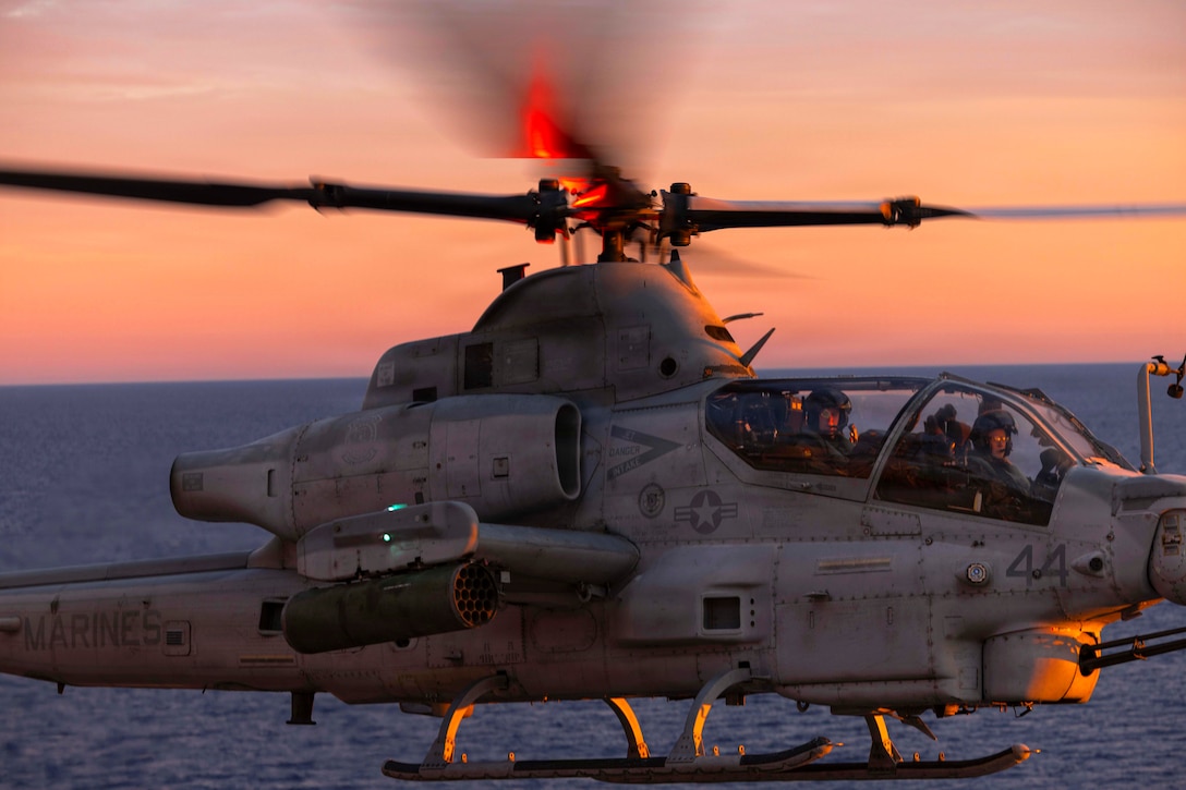 Two people wearing flight gear sit in a helicopter, while landing on a ship at sea that is not in frame.