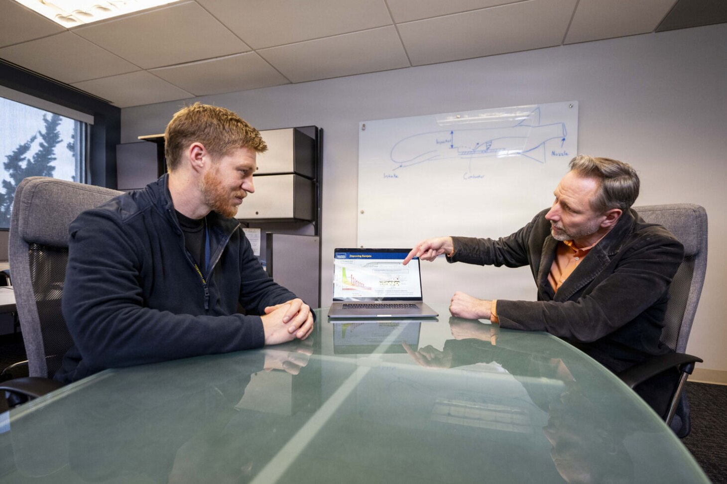 Two men wearing civilian attire sit at a conference table. The man to the right points at a laptop on the table as the other man looks on.