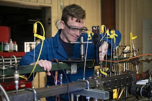 A man dressed in a lab coat and goggles, holds a metal bar on a device that has other metal bars and wires protruding from it.