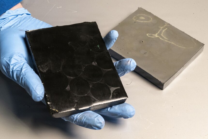 A person holds a black block in a gloved hand. A gray block is on a table nearby.