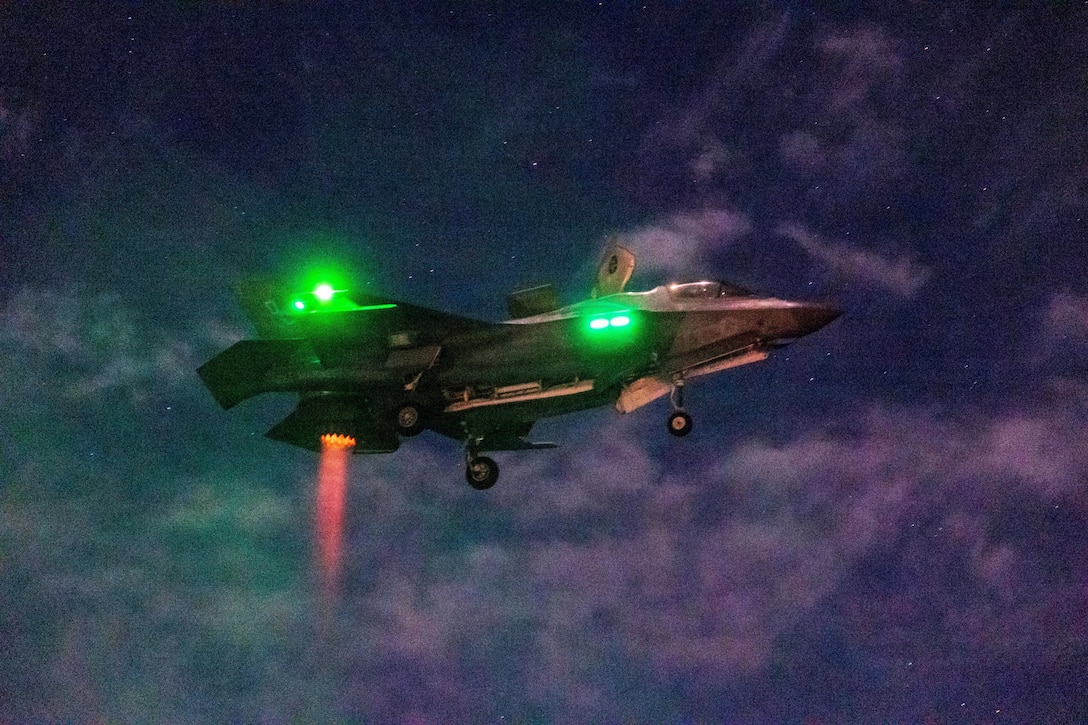A plane flies through the night sky with green glowing lights on the right side and red exhaust fumes glowing below the aircraft.