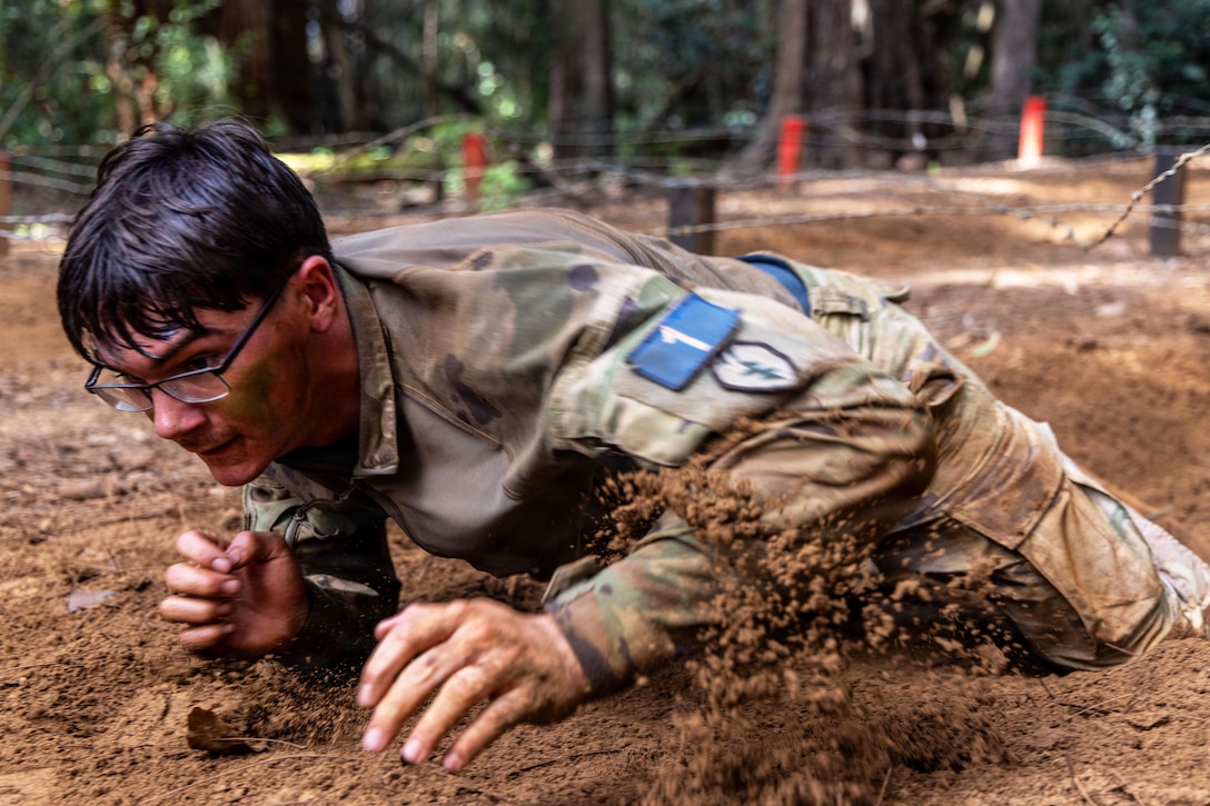 A man wearing a camouflage military uniform and glasses crawls on his hands and knees through a muddy obstacle course.
