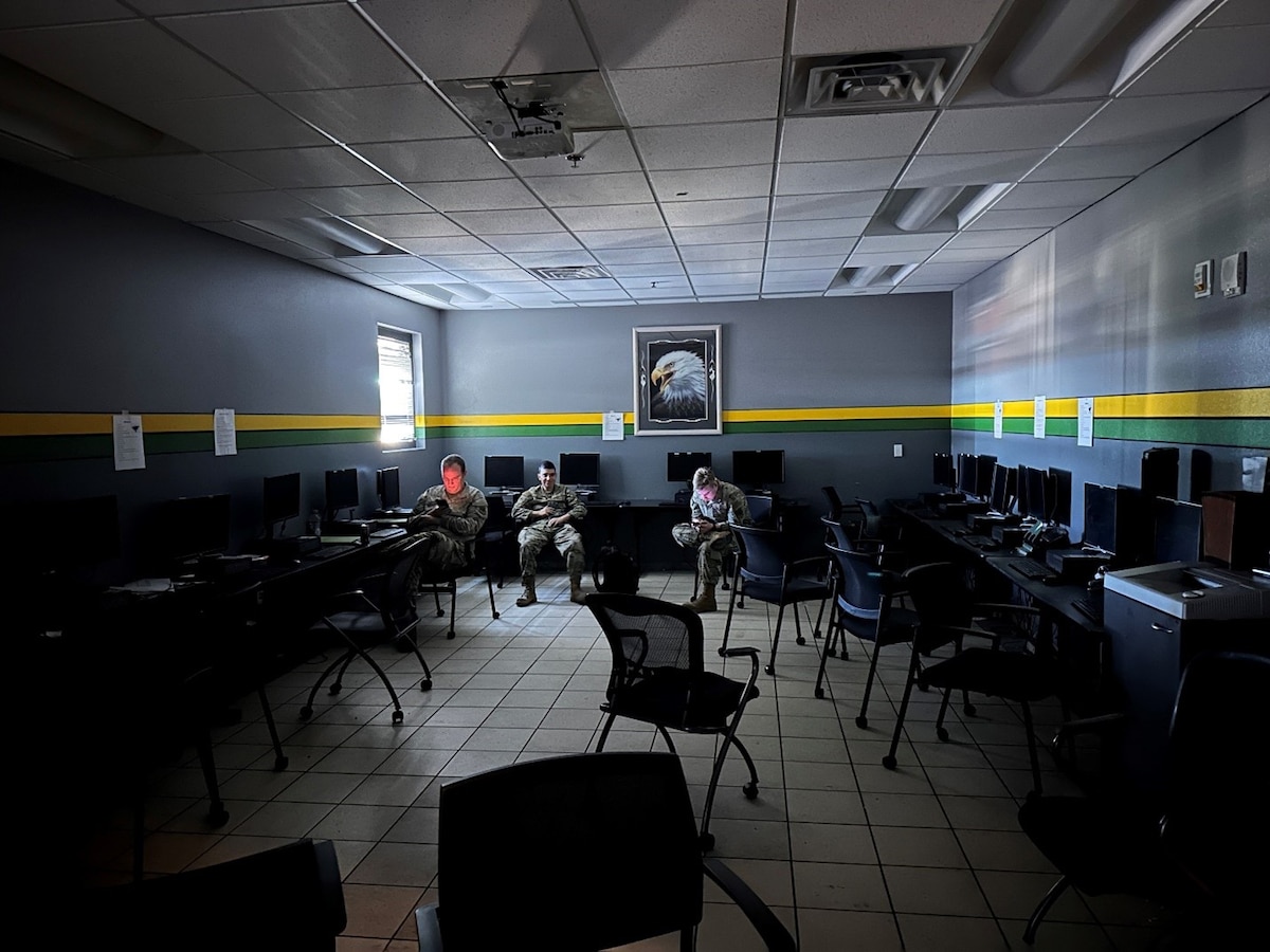 Man in Air Force uniform sits in dark room working on a laptop. Floor is grey tile. Photo of eagle on wall.