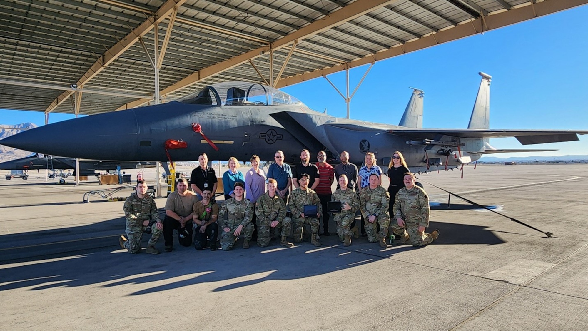 Group of individuals stand in front of Air Force fighter on tarmac. Day is sunny.