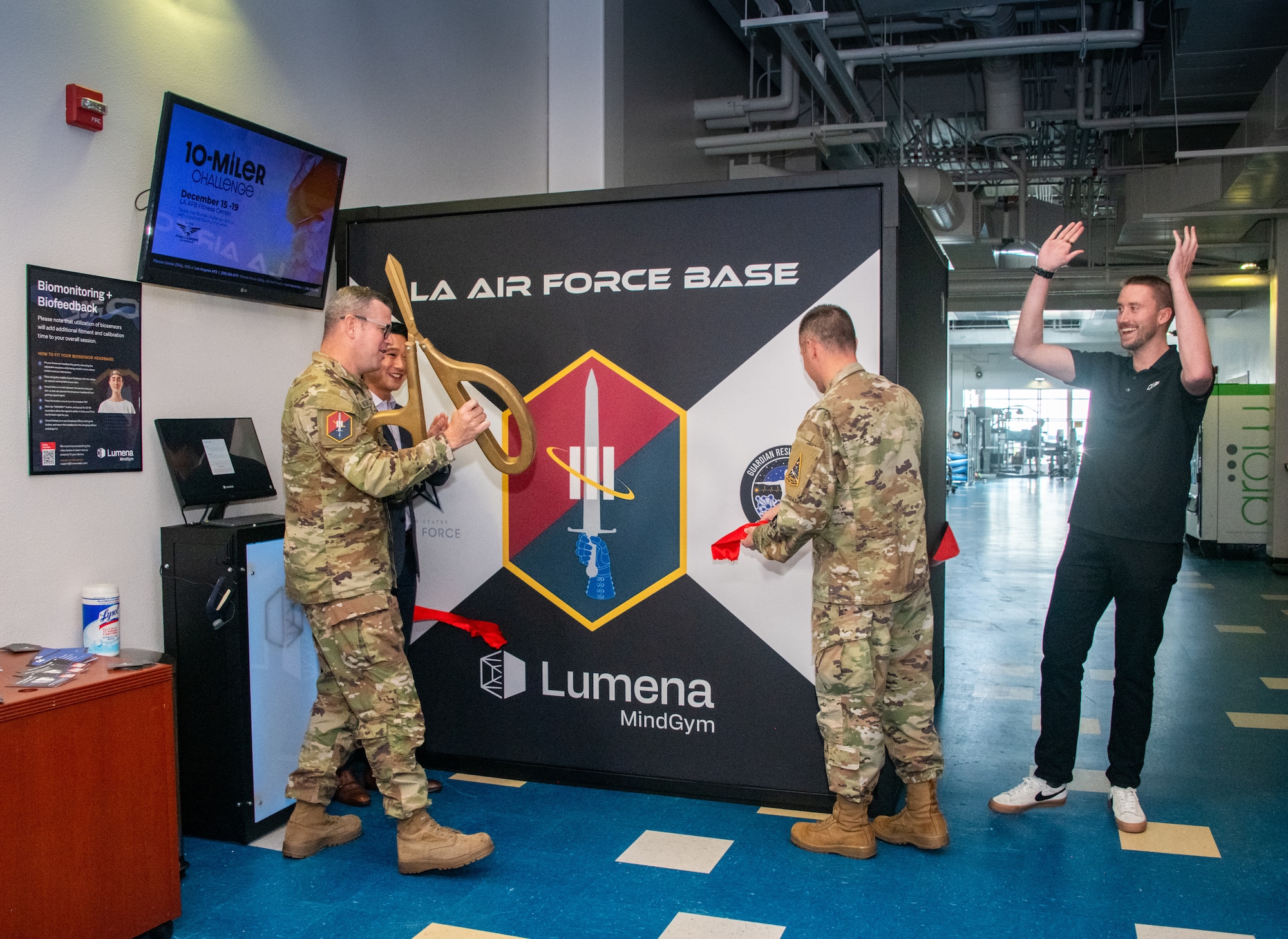 Two men in military OCPs cut a red ribbon around a nine-foot squared box known as the MindGym. Another man, the Lumena representative, raises his hands in excitement at the ribbon cutting and grand opening of the MindGym.