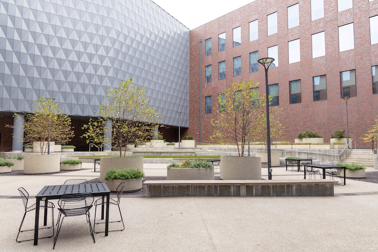 A three story building with grey exterior on the left side and red brick exterior on the right side. Concrete in the foreground with planters, trees, and tables and chairs.