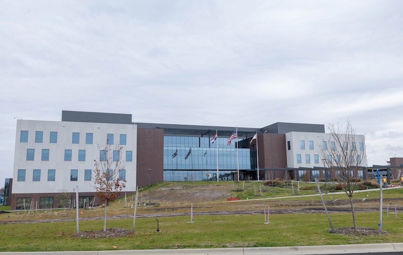 A beige and brown brick building with three stories and glass windows in the front sits on a green grassy hill with two flag poles in the front.