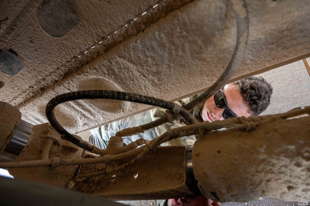 An airman wearing sunglasses works on an aircraft’s mechanics as seen through a small opening.