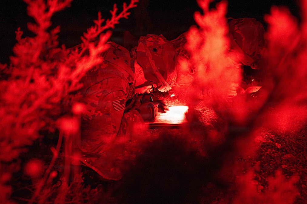 A soldier in tactical gear lies in a dark field illuminated by red light while writing on a map as seen through bushes in the foreground.