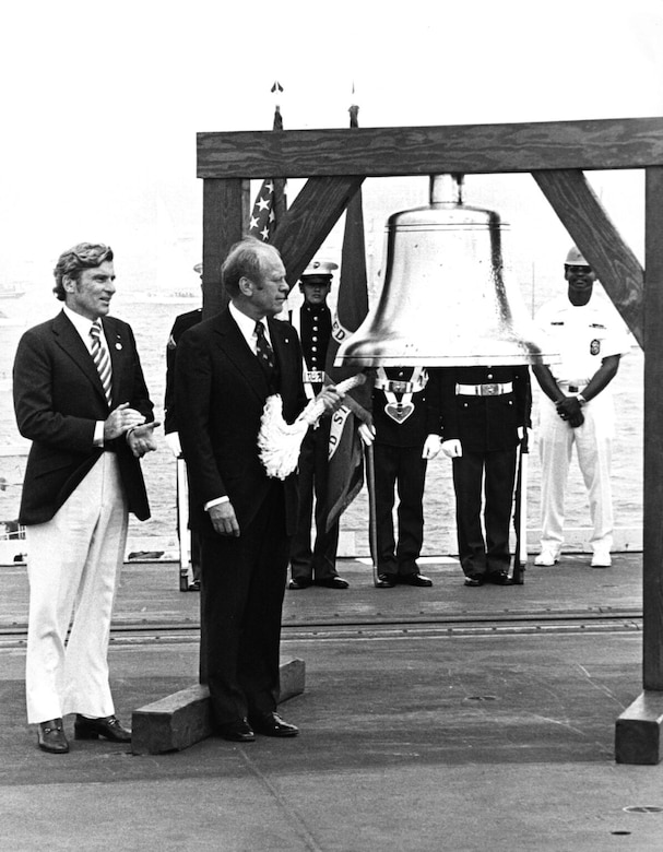A man in a suit rings a large ceremonial bell on a ship as another man in similar attire stands beside him. There are four people in military dress uniforms standing in the background.