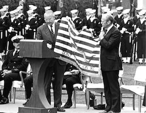 Two men in suits hold an American "Don't Tread on Me" flag, as people in military dress uniforms stand in formation.