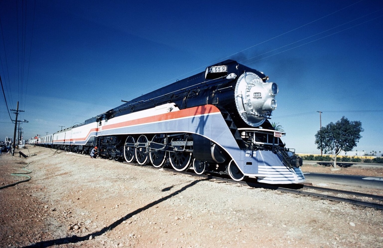 A steam locomotive painted in red, white and blue pulls a passenger train through a flat landscape under a blue sky.