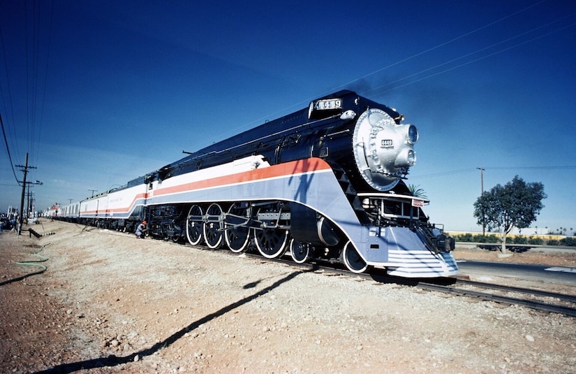 A steam locomotive painted in red, white and blue pulls a passenger train through a flat landscape under a blue sky.