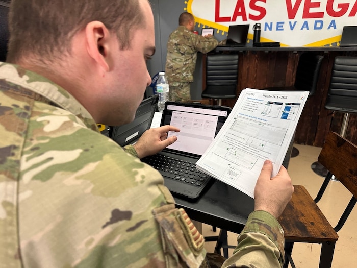 Man in Air Force uniform sits at computer and holds a piece of paper.