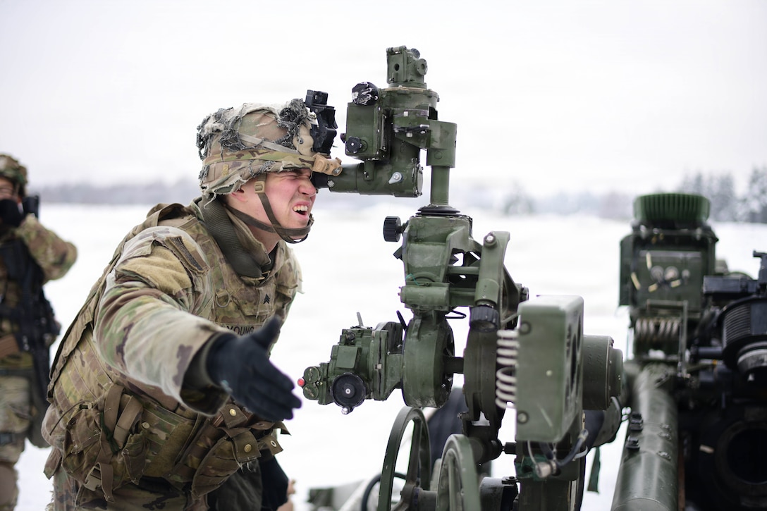 A soldier wearing a winter camouflage uniform looks through the sighting mechanism of an M777 howitzer positioned in snowy terrain.