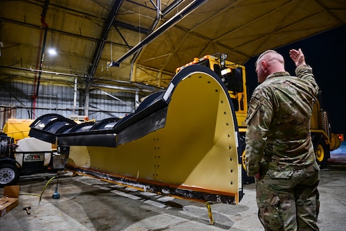 A man in a military uniform is waving a giant yellow vehicle into a large hangar.