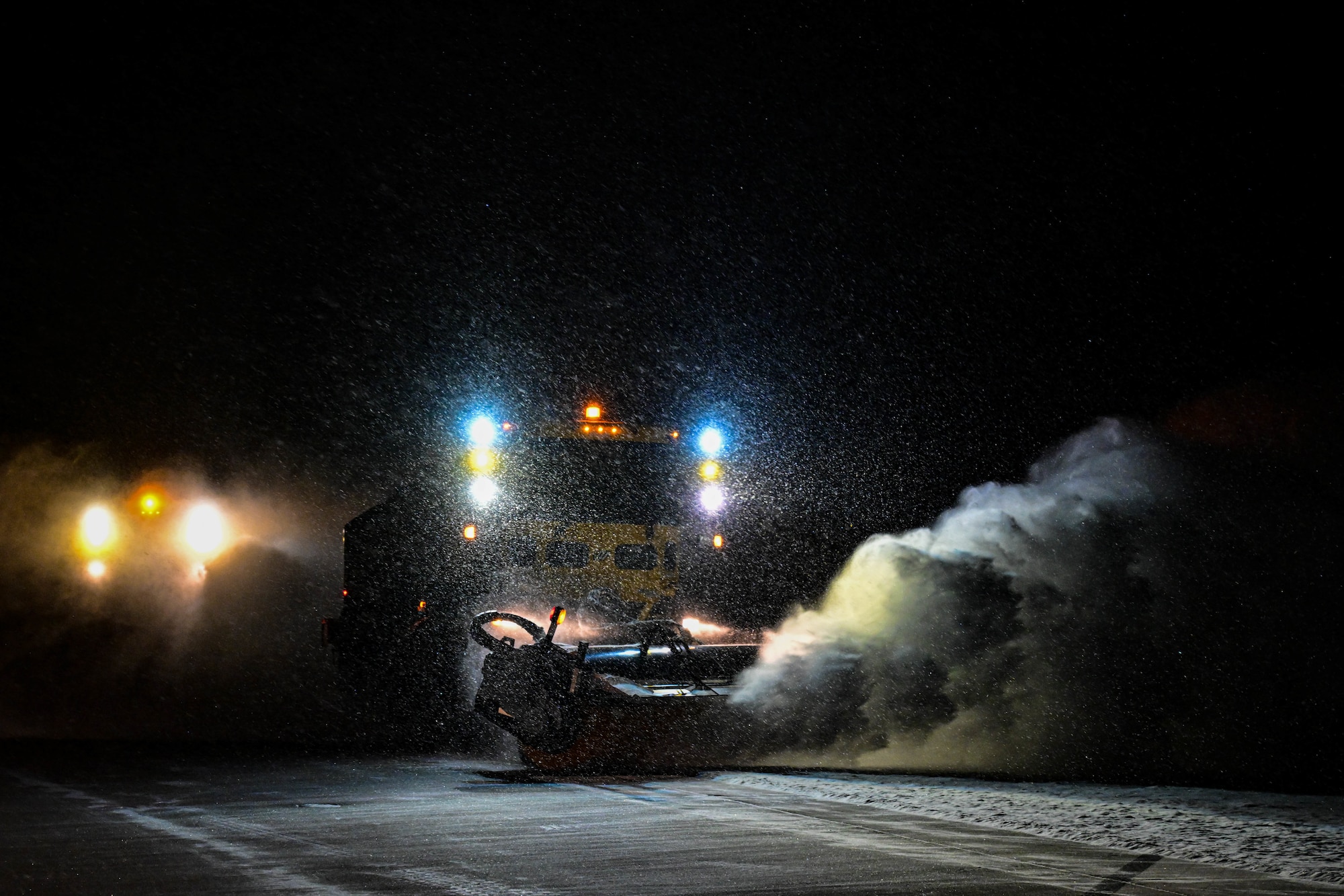A giant yellow vehicle driving in the dark, pushing snow and dust off to the side.