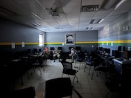 Man in Air Force uniform sits in dark room working on a laptop. Floor is grey tile. Photo of eagle on wall.