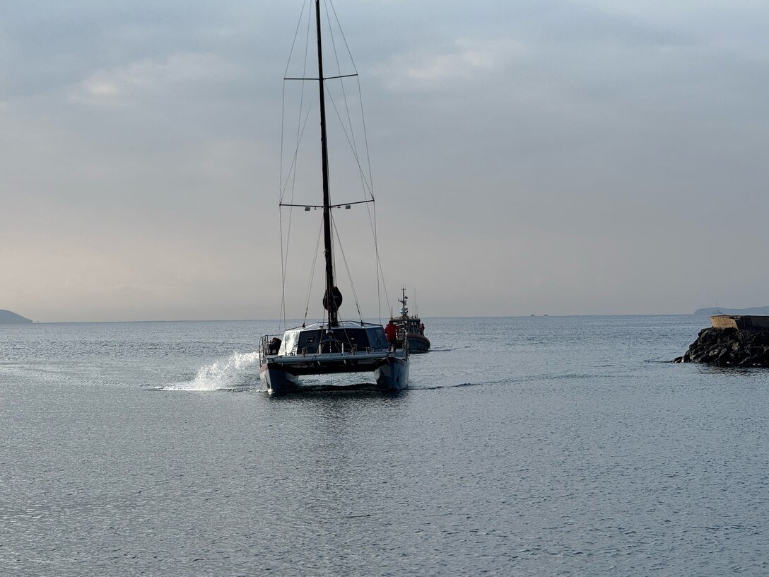A Coast Guard 45-foot Response Boat Medium and a sailing vessel move towards land. The water is still like glass and a grey hazy loom over the background.