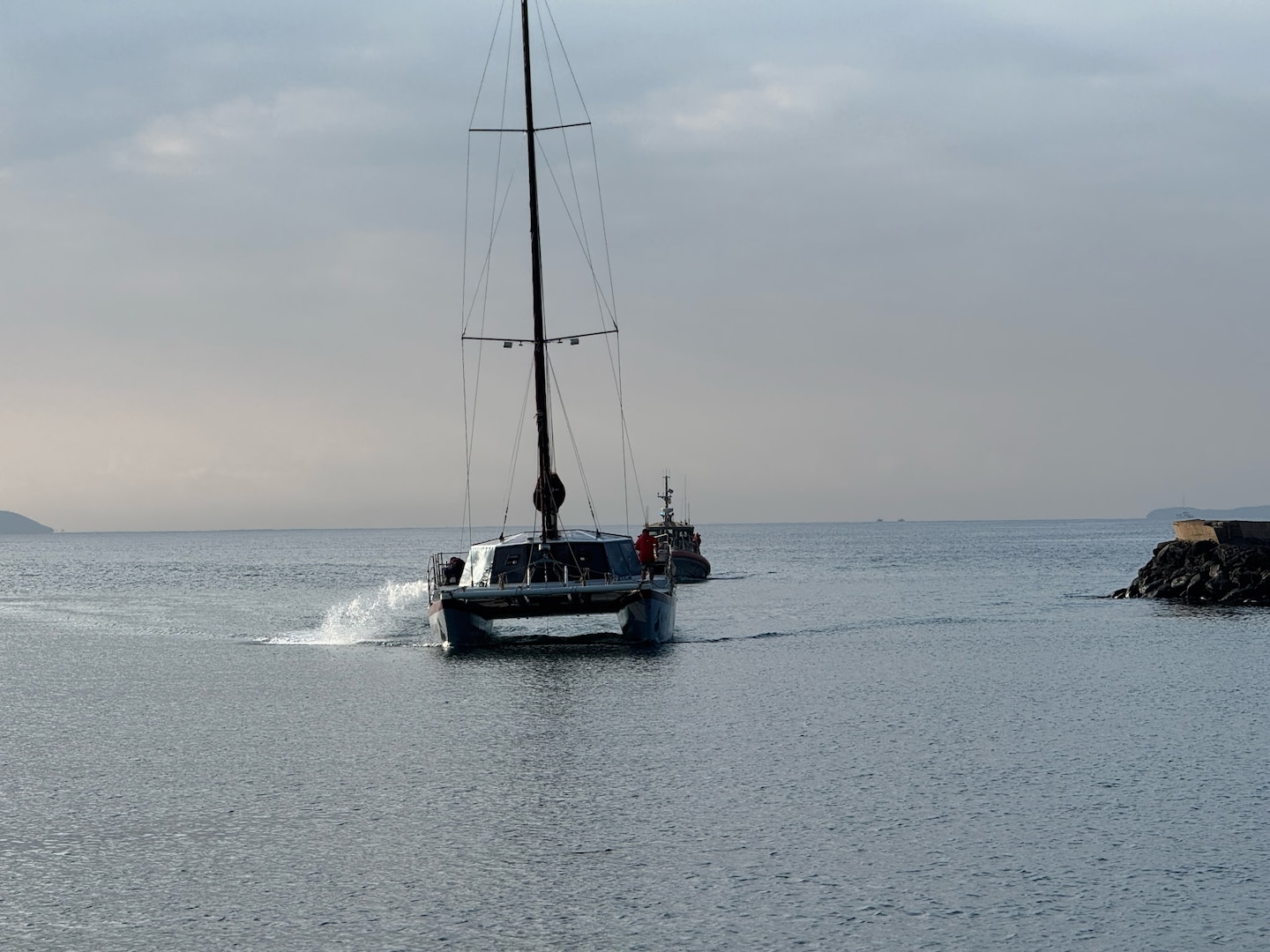 A Coast Guard 45-foot Response Boat Medium and a sailing vessel move towards land. The water is still like glass and a grey hazy loom over the background.