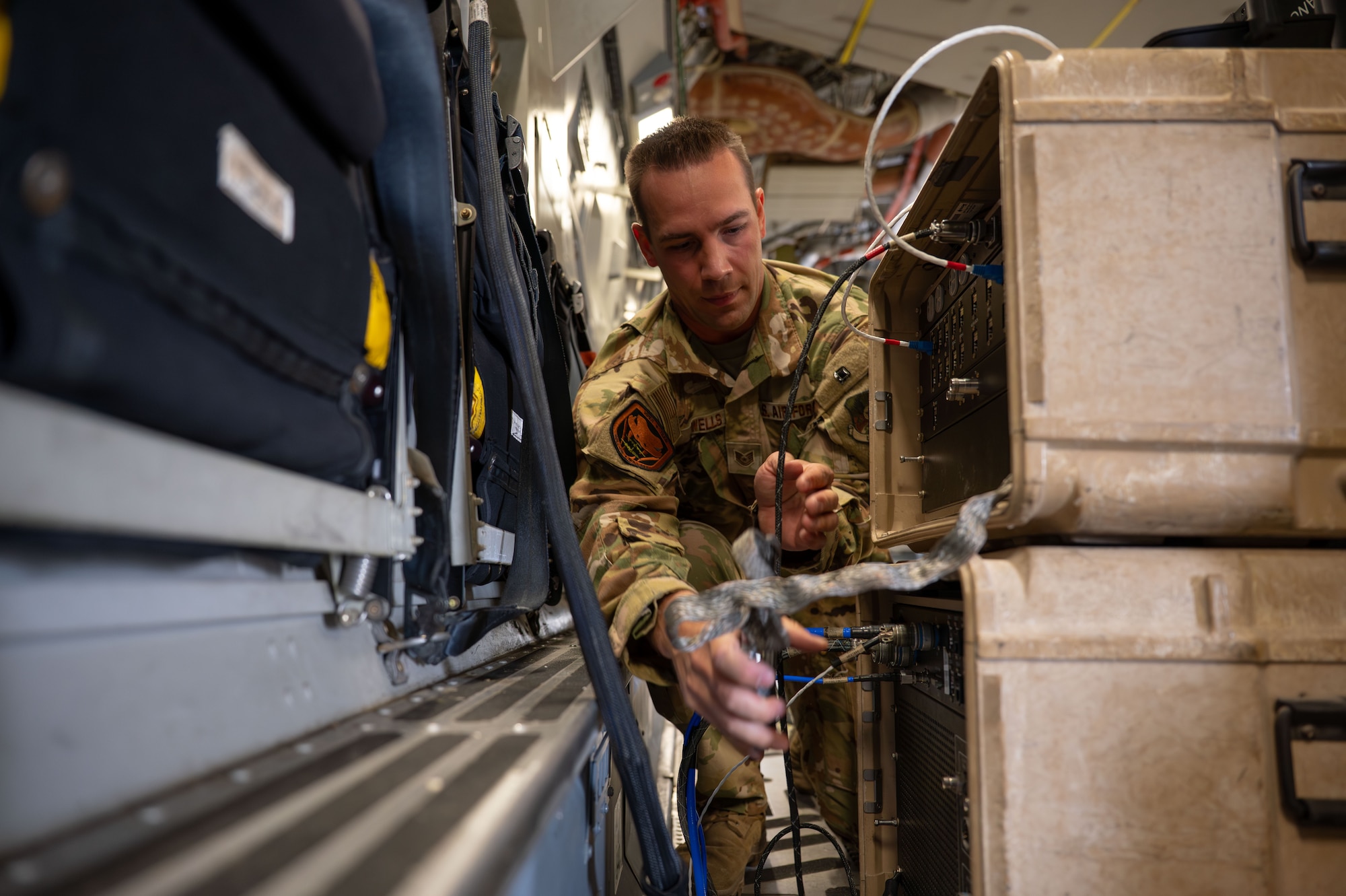 An Airman reaches between the wall of a C-17 and two cases with communications equipment stacked on top of each other to plug in a cable