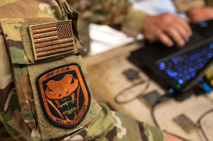 Close up of an Airman's right sleeve bearing a U.S. Flag patch and the Viper Team patch which has a snake in the middle with A C-17 and C-130 Aircraft. The bottom of the patch says "Roll-on, roll-off" There are hands typing on a laptop in the background