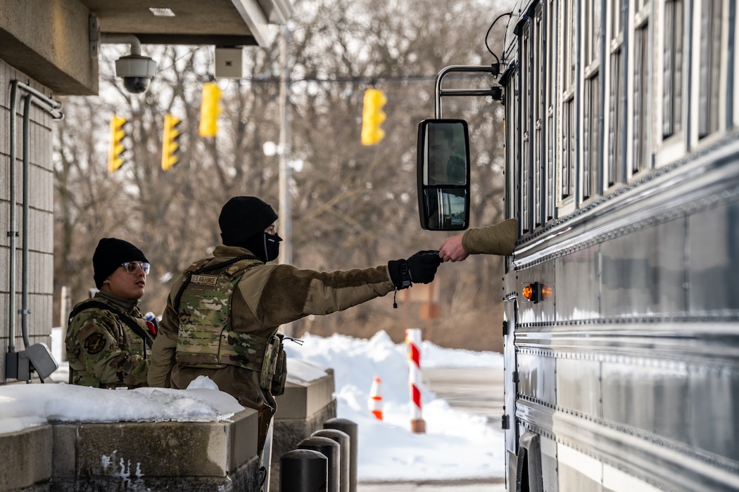 Two young men are wearing camouflage military uniforms, winter caps, and face coverings. One hands an ID card back to a bus driver.