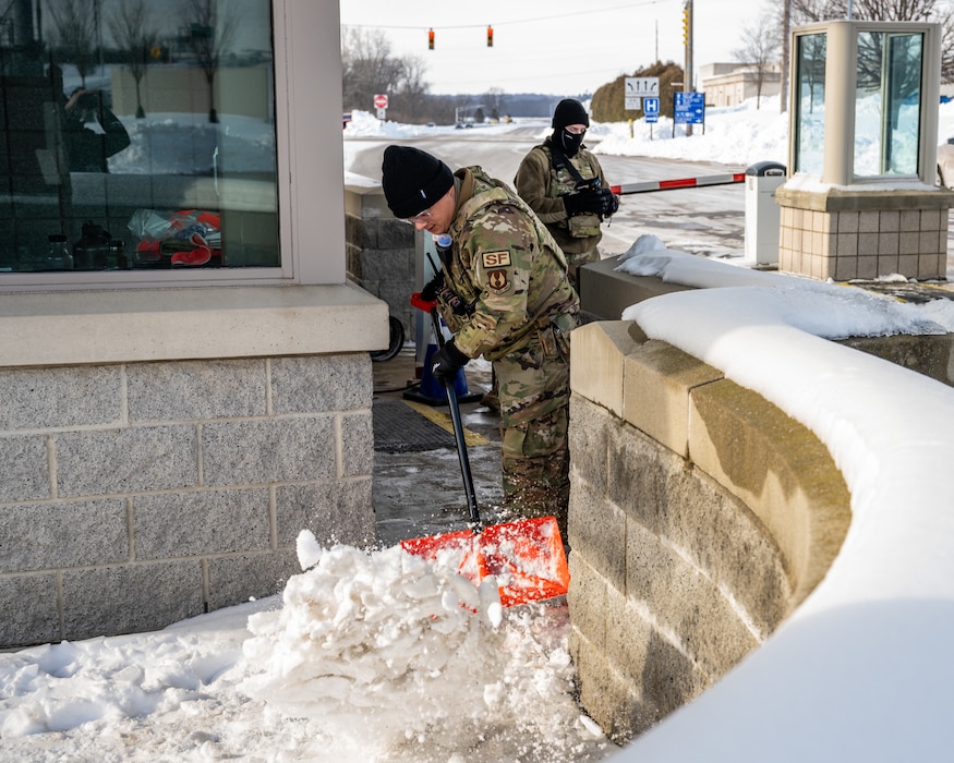 A young man, wearing camouflage military uniform and winter cap, uses an orange shovel to remove snow in the foreground