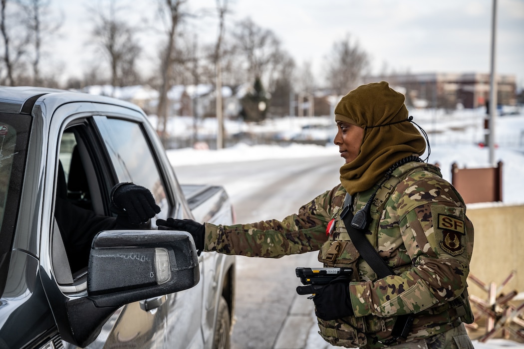 A young woman, wearing camouflage military uniform and brown head covering, hands an ID card back to a driver. Snowy landscape is out of focus in the background