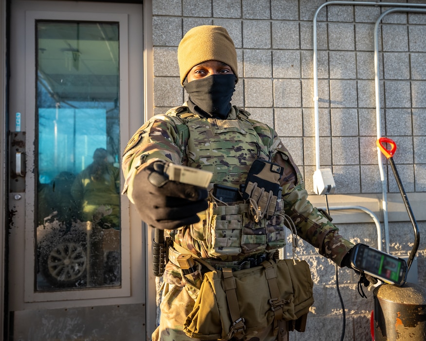 A young man, wearing camouflage military uniform, winter cap, and face covering, extends a hand holding an ID card forward towards view point
