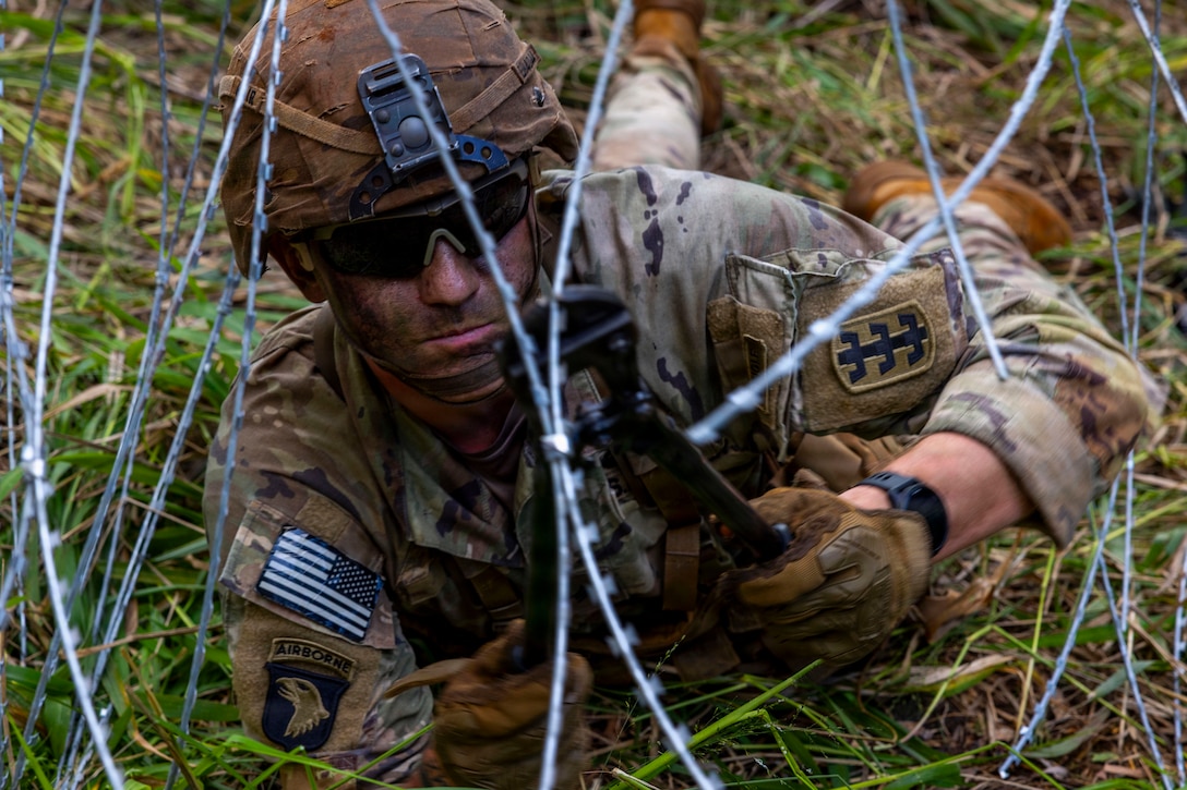 A man wearing a camouflage military uniform, helmet and dark glasses lies on his stomach in a grassy area to cut a wire.